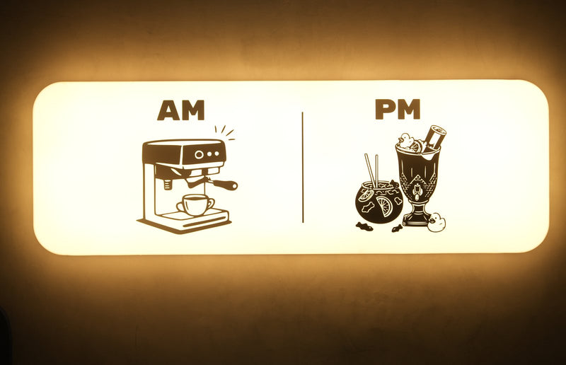 A lit sign split in two: AM above a coffee machine, PM above cocktails.