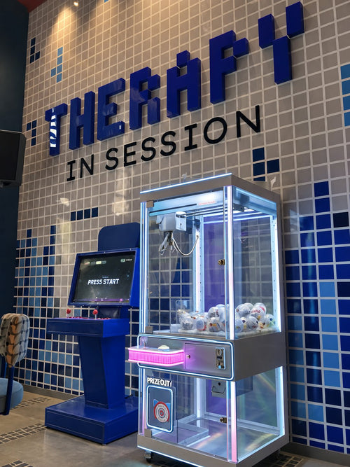 A claw machine and an arcade cabinet under a tiled wall that reads THERAPY IN SESSION.