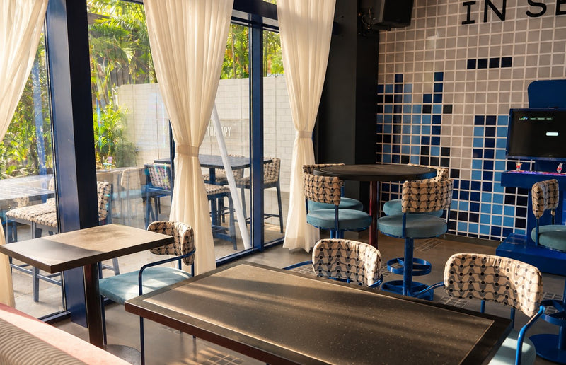 Tables and patterned chairs beside floor-to-ceiling windows and white curtains, with a blue tiled wall behind.