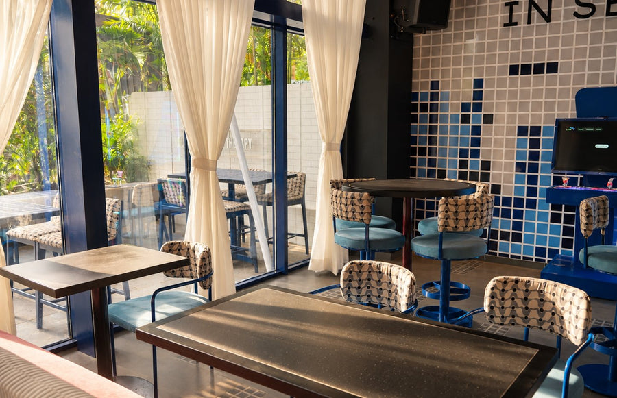 Tables and patterned chairs beside tall windows and white curtains.