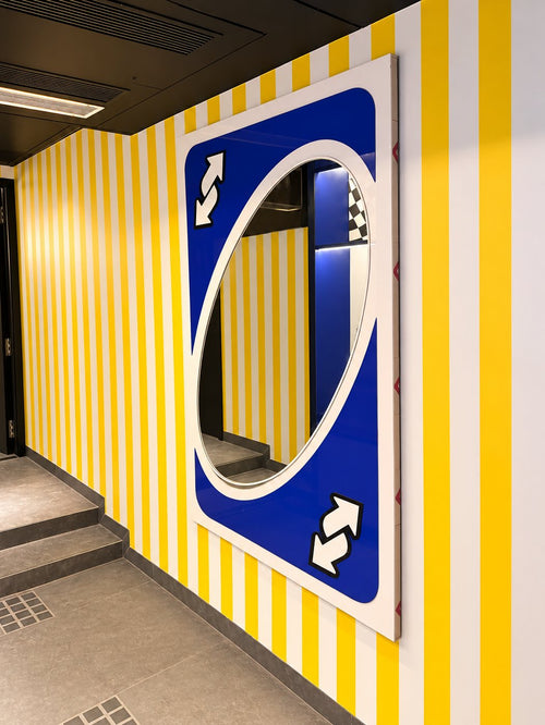 A mirror shaped like a giant blue UNO reverse card on a yellow and white striped wall.